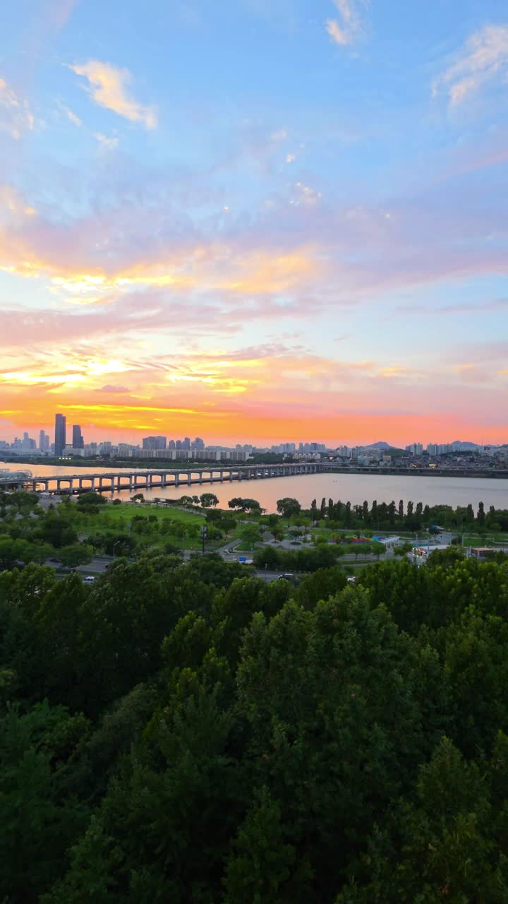 A beautiful vertical aerial shot pans left to reveal a spectacular golden hour sunset over the Seoul cityscape, featuring the lush Hangang Park, the Han River, and the iconic Banpo Bridge in summer