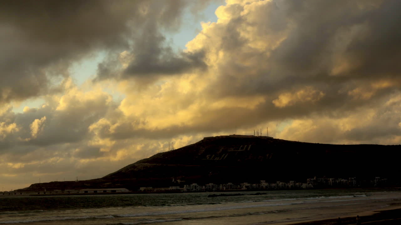 Golden clouds drift over a coastal mountain marked with bold Arabic letters as waves gently break along the quiet shoreline.