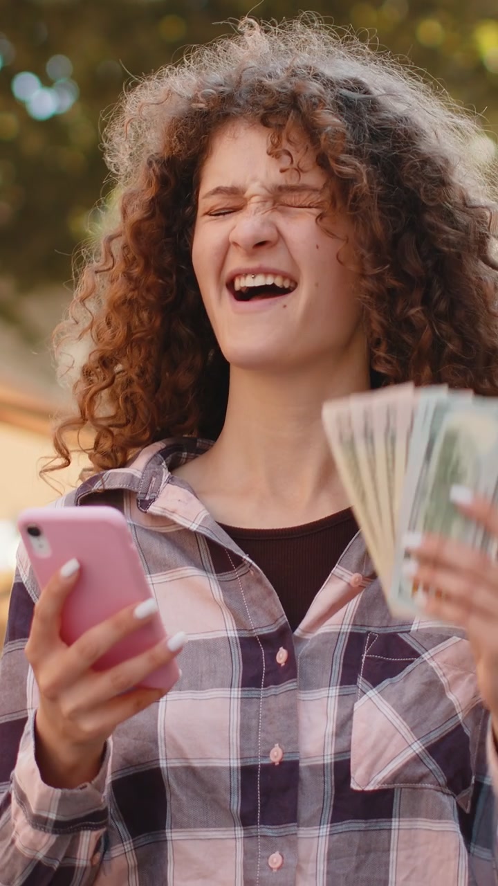 Happy young woman counting money dollar cash use smartphone calculator app in urban city street