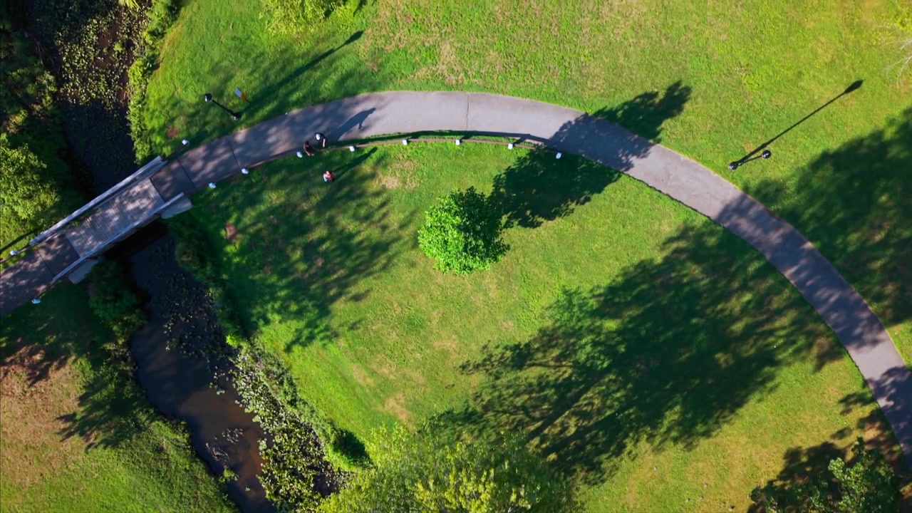 Aerial top down view of a winding park pathway crossing a creek under a small bridge, showing greenery and the peaceful outdoors on a sunny day. The camera is tilted straight down and dolly in slowly.