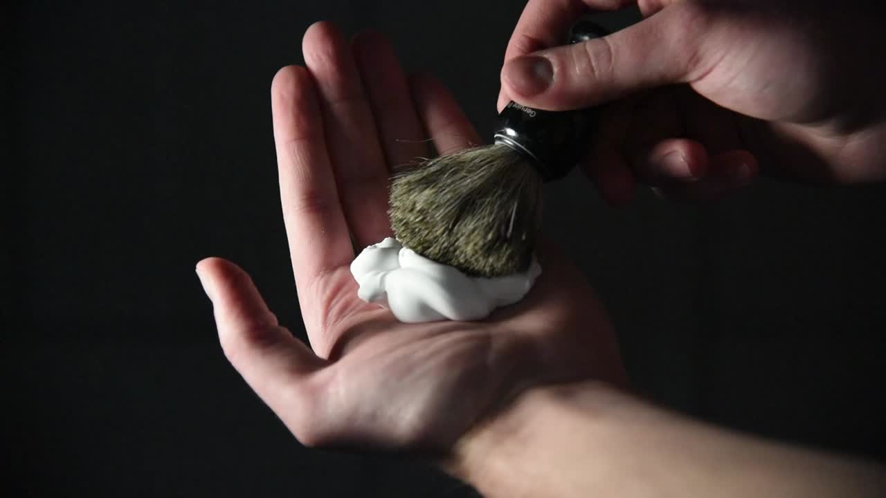 Close up of Caucasian hand with shaving foam in center. Lathers black shaving brush in the foam. Dark indoor background