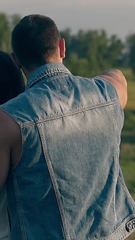 wonderful close backside view young couple stands against green trees on meadow and man raises hand slow motion