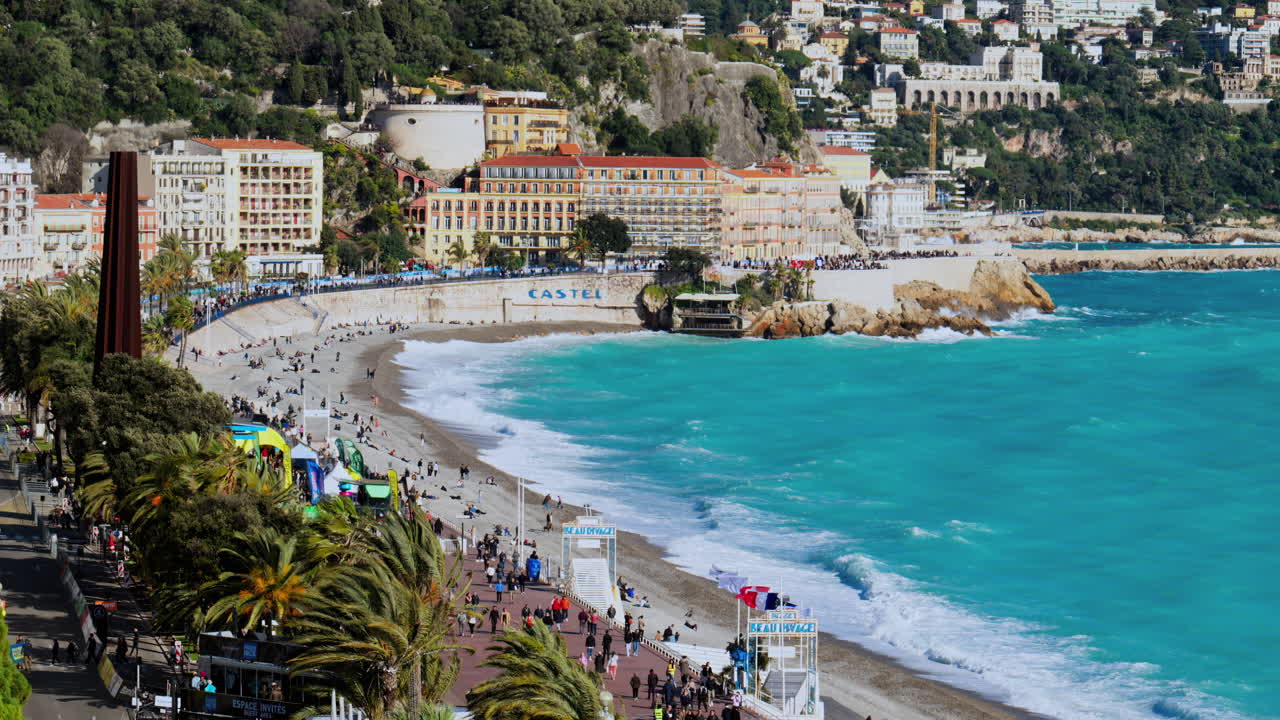 Nice, France - March 17, 2025: Distant view of people walking on the coast of the city with water crashing on the rocks and the buildings on the background