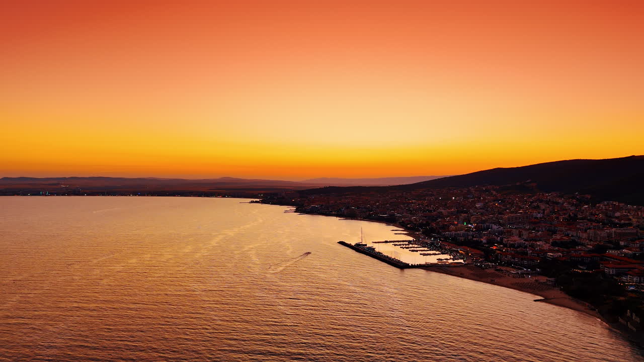 Pôr do sol na costa com vista para o litoral e pequenos barcos. O sol se põe sobre a água, lançando um brilho alaranjado sobre o litoral e o porto com barcos.