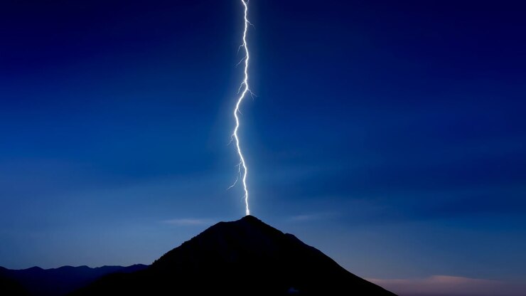 Un fulmine bianco-blu brillante balena da un cielo scuro e colpisce la cima di una montagna, creando un bagliore intenso, scariche frastagliate, un contrasto netto e un'illuminazione drammatica su un terreno alpino remoto al crepuscolo.