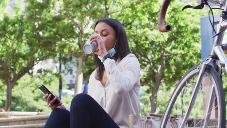 mujer afroamericana con máscara facial usando un teléfono inteligente bebiendo café en un parque de la ciudad