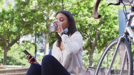 african american woman wearing face mask using smartphone drinking coffee in city park