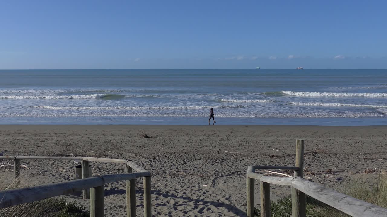 Person walking in wet sand on incoming tide as ships anchor off-shore