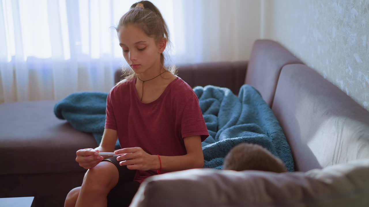 Girl with ponytail in maroon shirt checks thermometer reading while sitting beside sick person covered in blanket showing concern care and responsibility in calm softly lit indoor living room setting