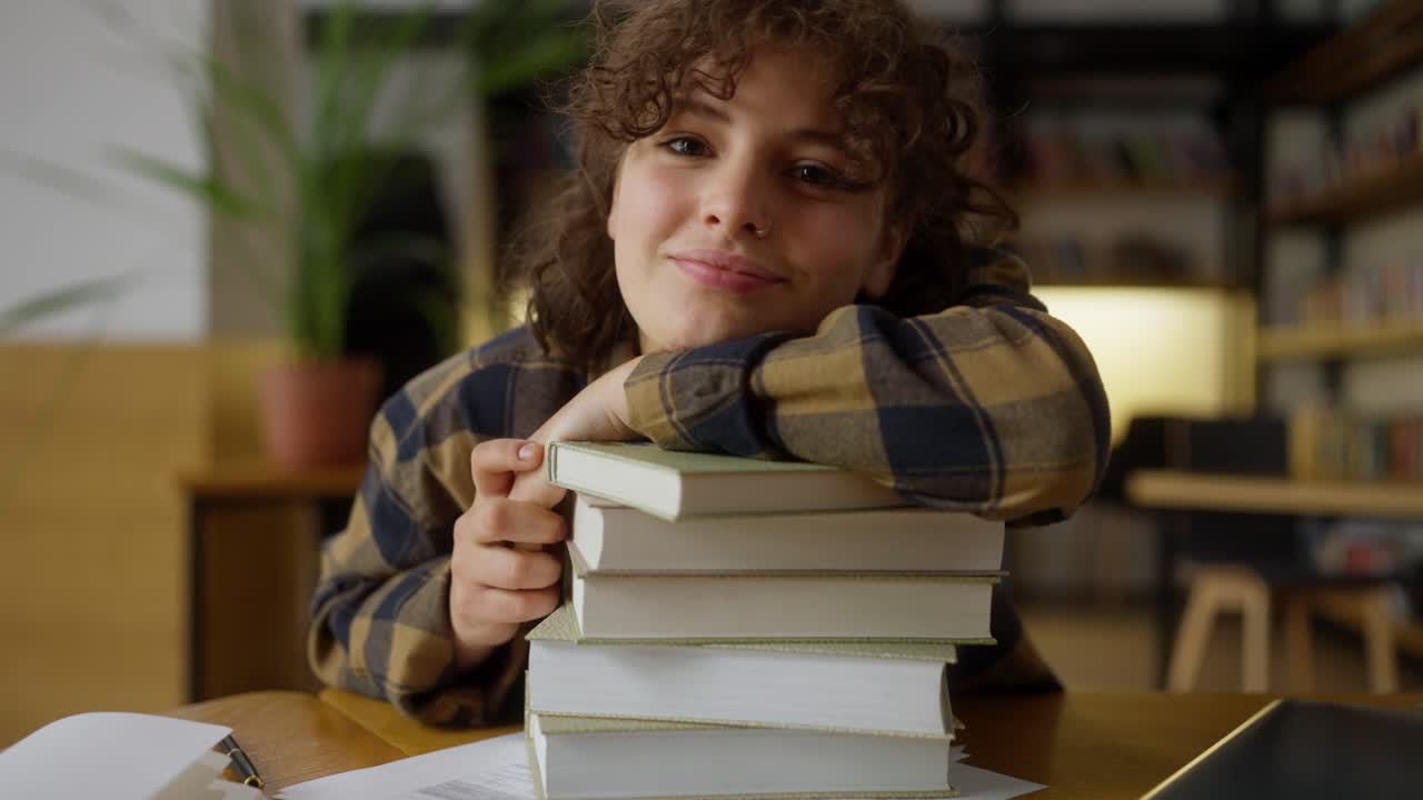 retrato de una estudiante feliz con el cabello rizado que sonríe y se apoya en una pila de libros en una mesa en la biblioteca