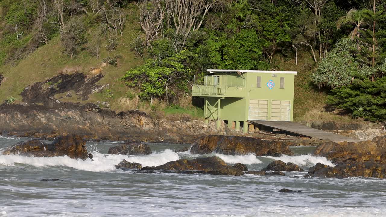 Waves crash against rocks near a green hut on a lush, forested coastline. Bright daylight enhances the vibrant scenery
