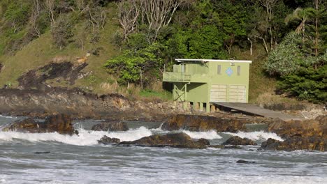 waves crash against rocks near a green hut on a lush, forested coastline. bright daylight enhances the vibrant scenery