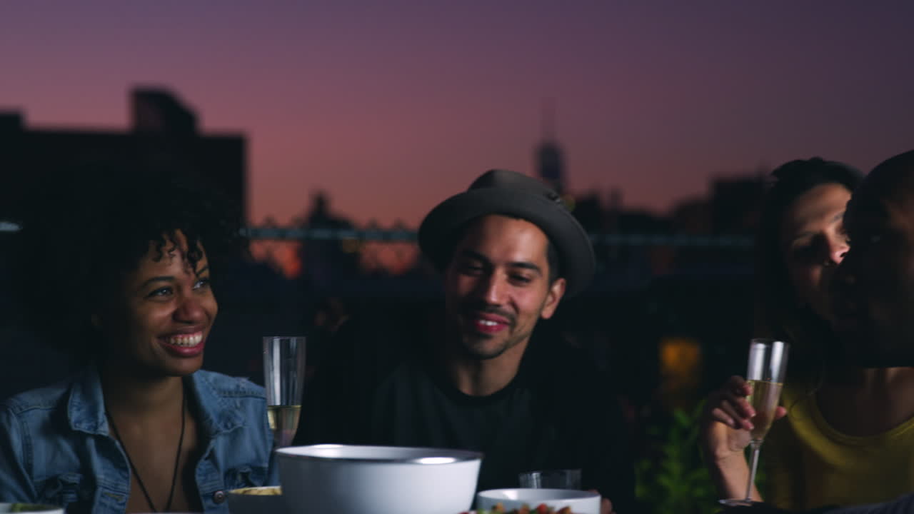 Amigos sentados ao redor de uma mesa em um terraço conversando ao anoitecer.
