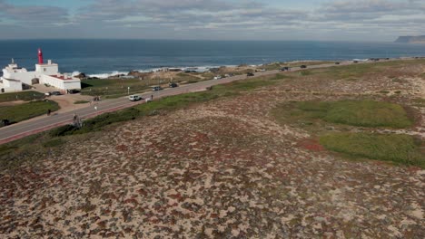 Vista-Aerea-De-La-Zona-De-Guincho,-Con-El-Faro-De-Cabo-Raso-Y-Algunos-Carros-Moviéndose-Sobre-Marginal