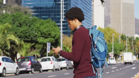 hombre afroamericano en la ciudad, usando un teléfono inteligente, con auriculares y mochila cruzando la calle