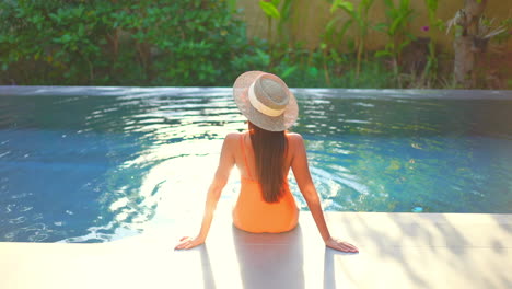 Vista-Trasera-De-Una-Mujer-Sentada-En-El-Borde-De-La-Piscina-Con-La-Luz-Del-Sol-Reflejada-En-El-Agua