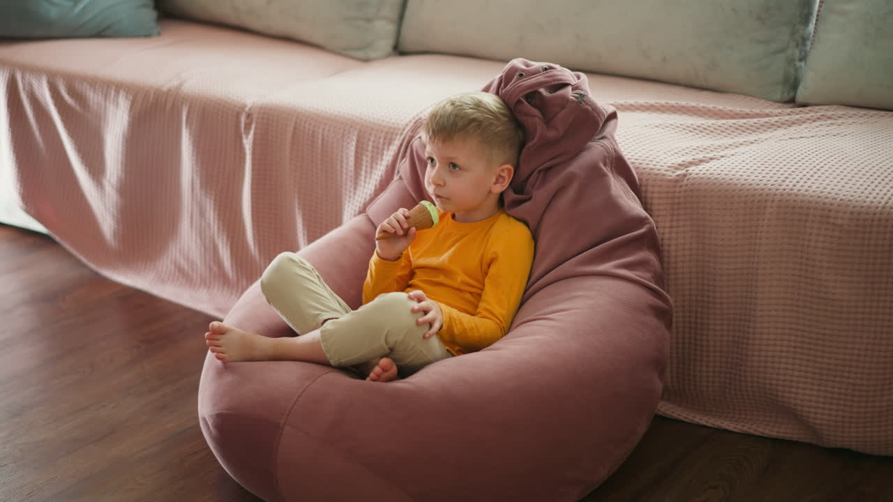 Little boy relaxing on large soft bean bag, happily eating mint chocolate ice cream cone indoors, wearing bright yellow shirt, legs crossed comfortably