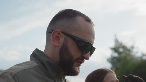 close up of man wearing dark sunglasses gazing upward beside woman against cloudy sky and blurred trees highlighting reflective lenses and contemplative expression in sunny outdoor setting