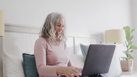 happy senior caucasian woman sitting on bed making video call on laptop and waving, slow motion