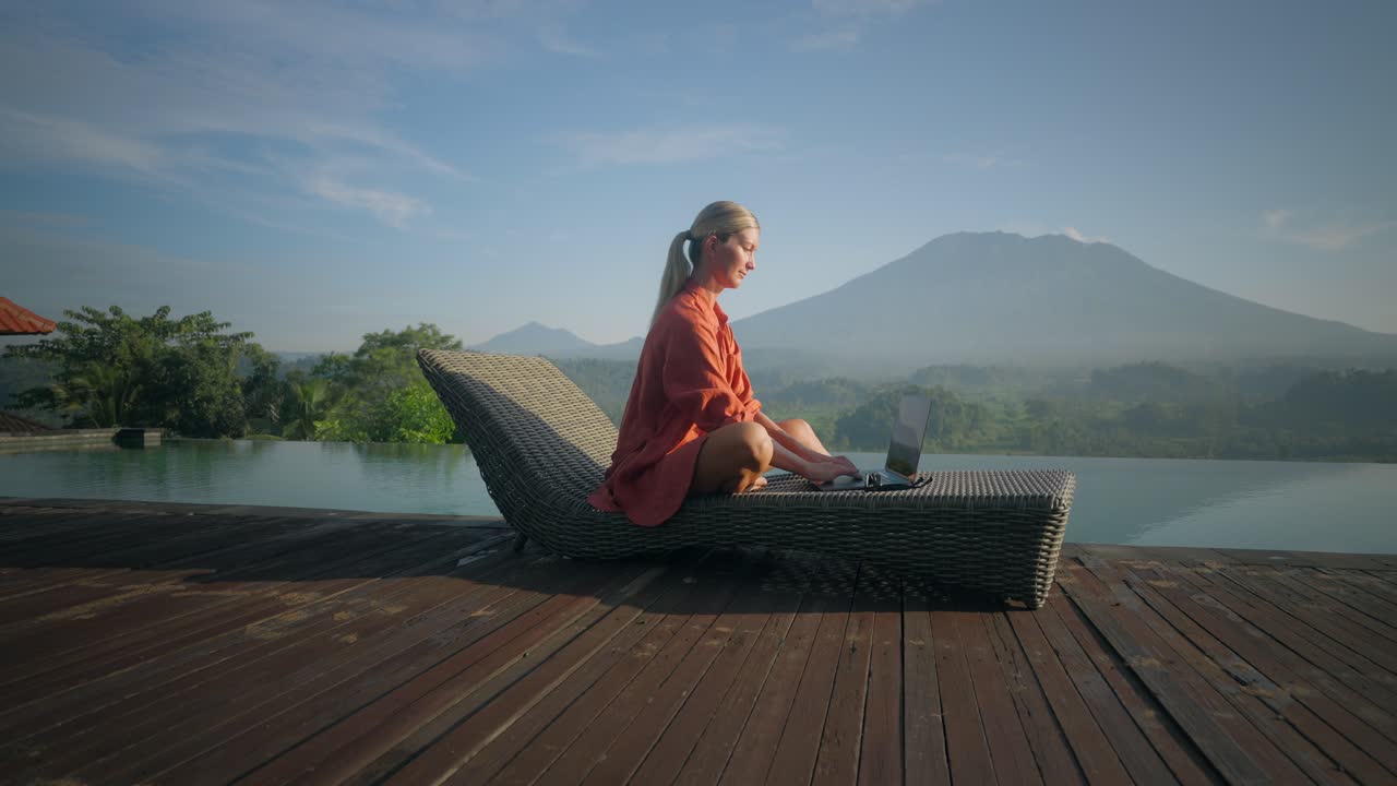 수영장 라운지 의자에 앉아 노트북에 입력하는 여성 디지털 유목민은 햇빛을 즐기는 선글라스를 착용합니다.