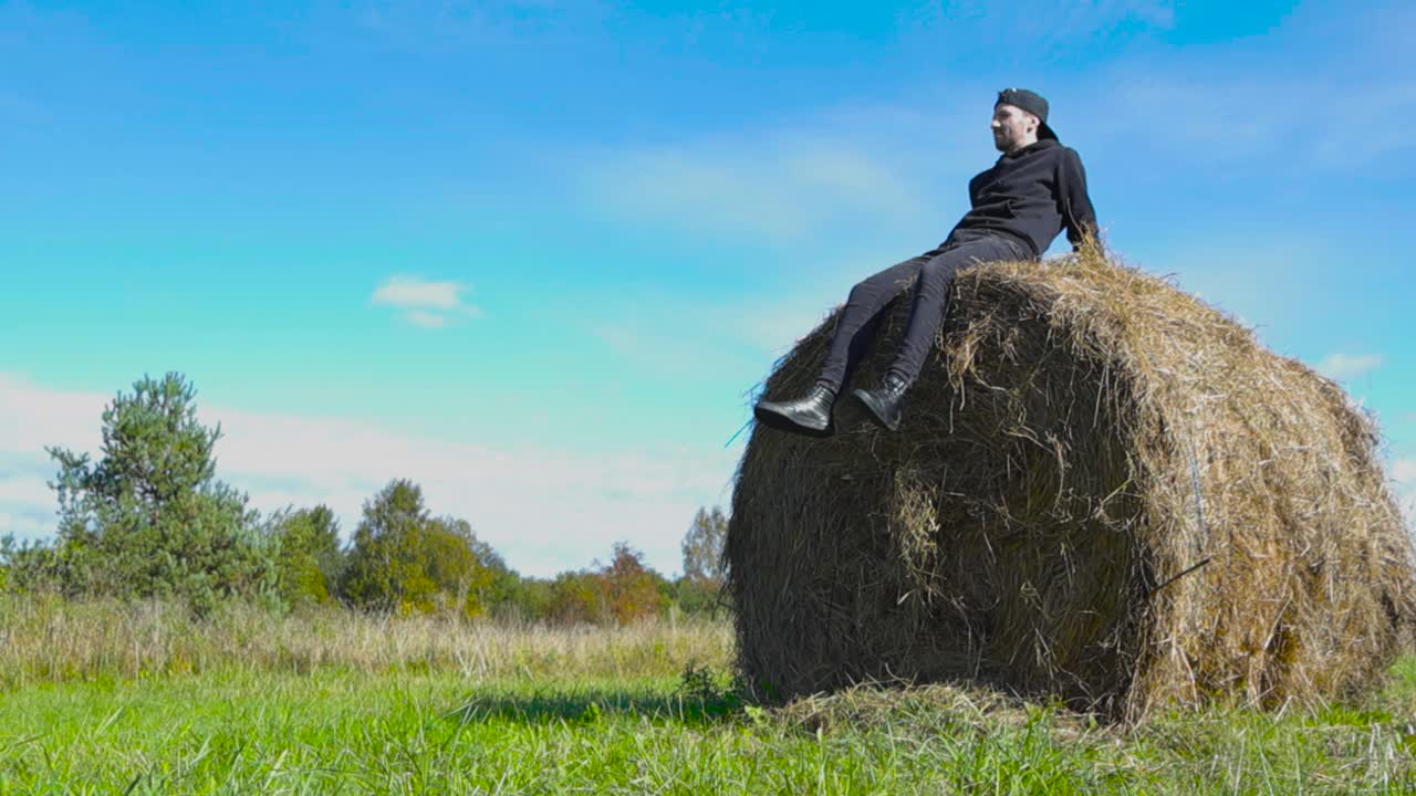 Caucasian male person sitting on top of a rounded hay bale during summer time while sun is shining and moves his legs in slow motion making hay fall down. Surrounded by farm field, green grass, nature