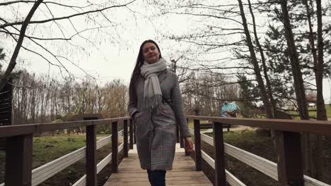 mujer joven con el abrigo gris está caminando sobre el puente de madera hacia la cámara