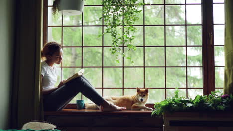 woman reading a book by the window with a dog
