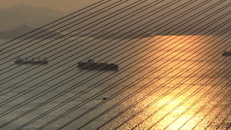 Aerial-drone-shot-of-ships-behind-vertical-suspenders-of-a-wide-suspension-bridge-during-golden-hour