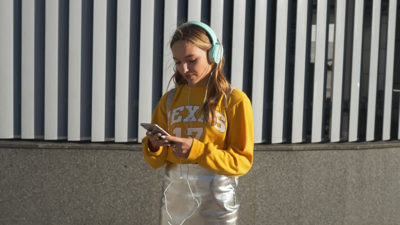 Ritratto di giovane ragazza carina e attraente su sfondo urbano che ascolta musica con le cuffie. Donna che indossa una blusa gialla e una gonna argentata.