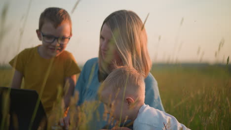 a woman in a blue gown, wearing a headset around her neck, sits in a grassy field working on a laptop as her two young sons happily watch her