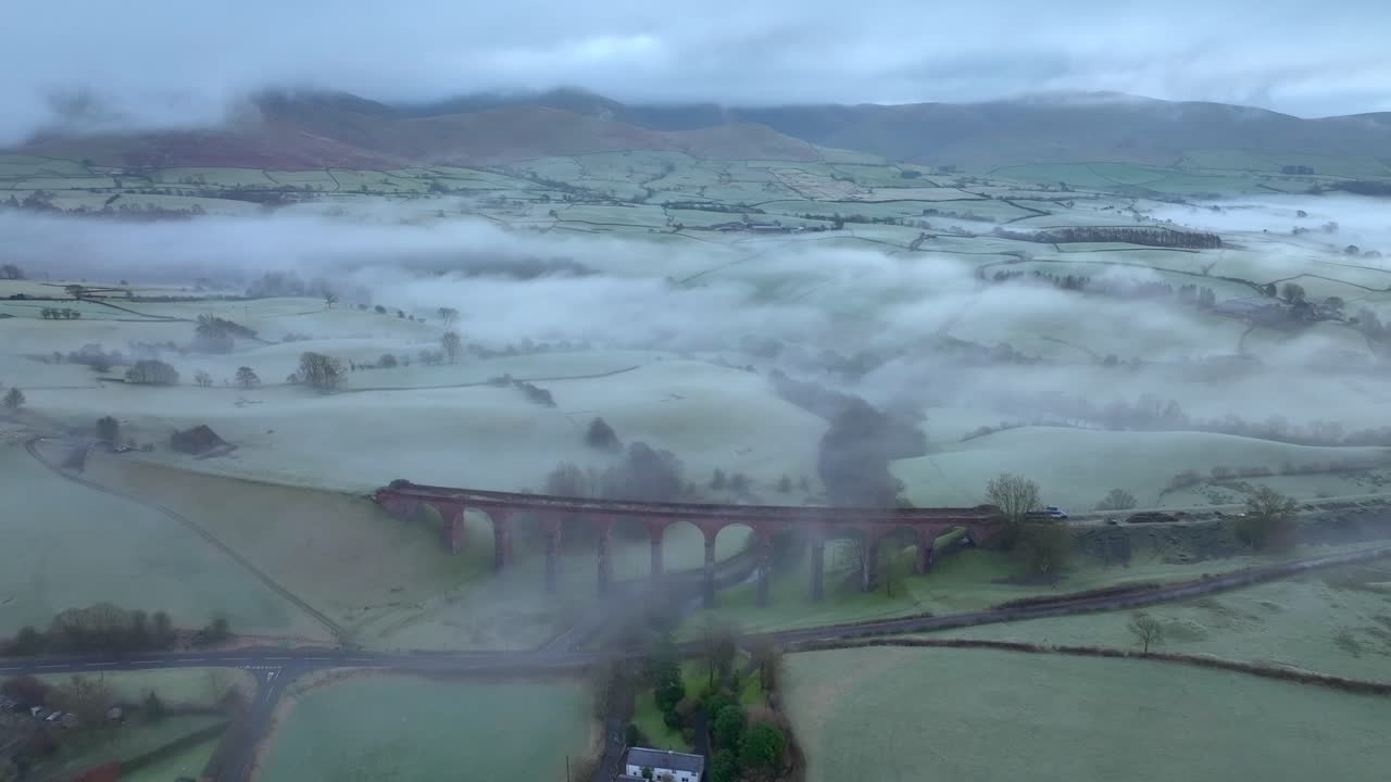 se acerca el puente de viaducto de piedra abandonado rodeado de tierras de cultivo y envuelto por la niebla fría y la niebla en la mañana de invierno con montañas cubiertas de nubes