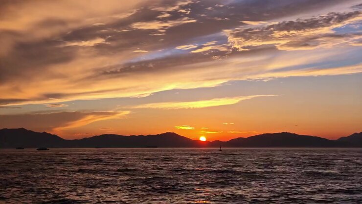 Golden hour sunset over ocean waves as boats and ships sail past distant mountain ranges under dramatic high-altitude clouds.