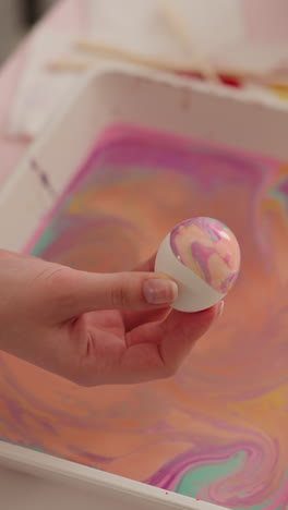 teenage girl holds easter egg covered with colorful patterns of ebru art over tray at table closeup slow motion. design and decor for holiday celebration