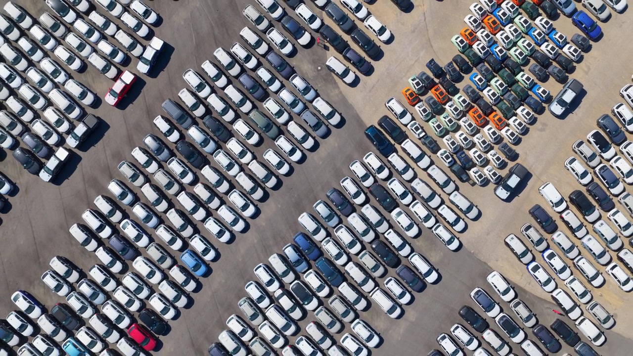 Droneoptagelse fra luften, der viser utallige splinternye biler pænt parkeret i rækker, klar til forsendelse eller salg ved et stort industrielt distributionscenter eller en fabriks parkeringsplads.