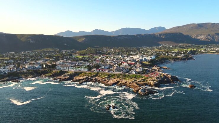 Suggestiva città costiera di Hermanus sulla Cape Whale Coast a Overstrand, che si affaccia sull'Oceano Atlantico con montagne sullo sfondo, ripresa aerea con drone in allontanamento