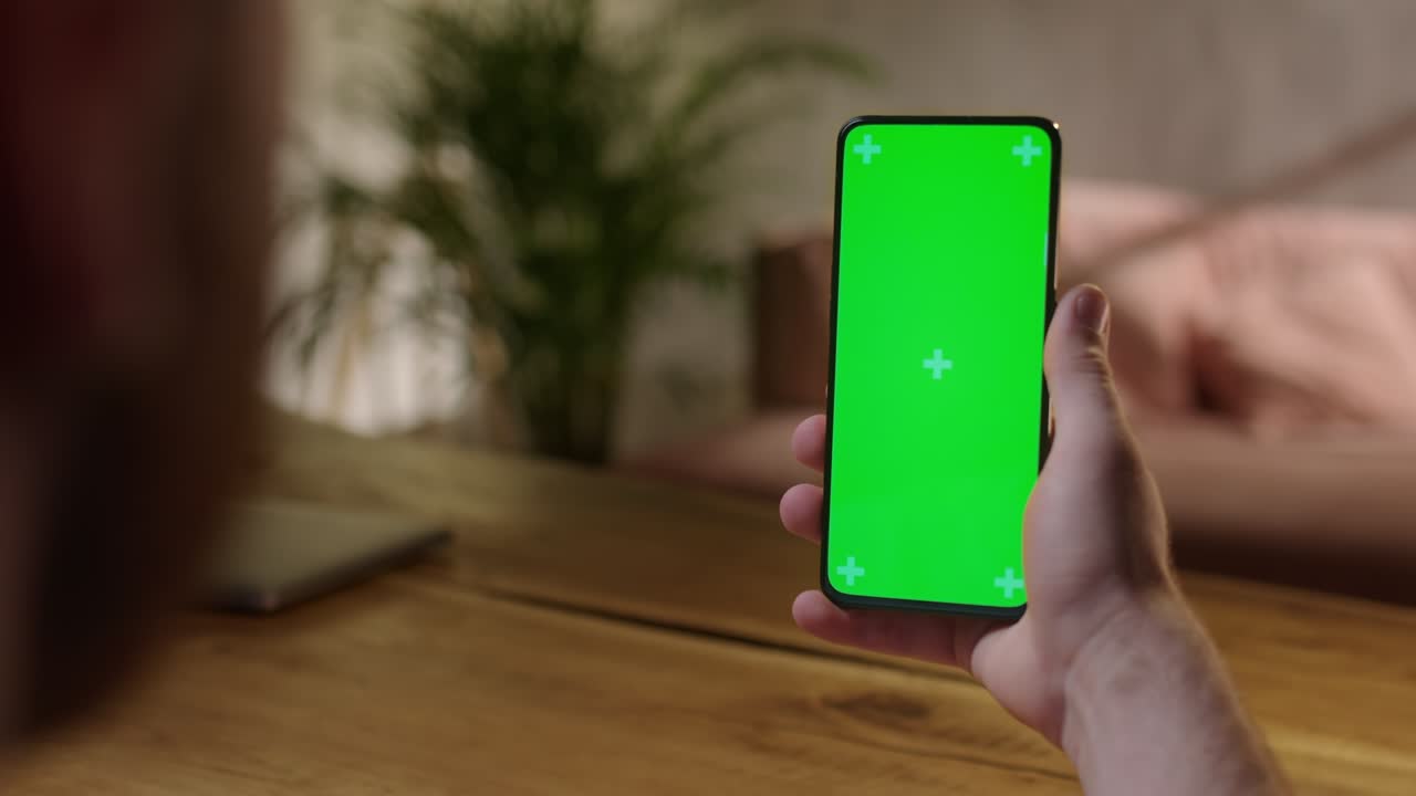 Handheld Camera: Young Man at Home Holding Chroma Key Green Screen Smartphone Watching Content Without Touching or Swiping. Browsing Internet, Watching Content, Videos
