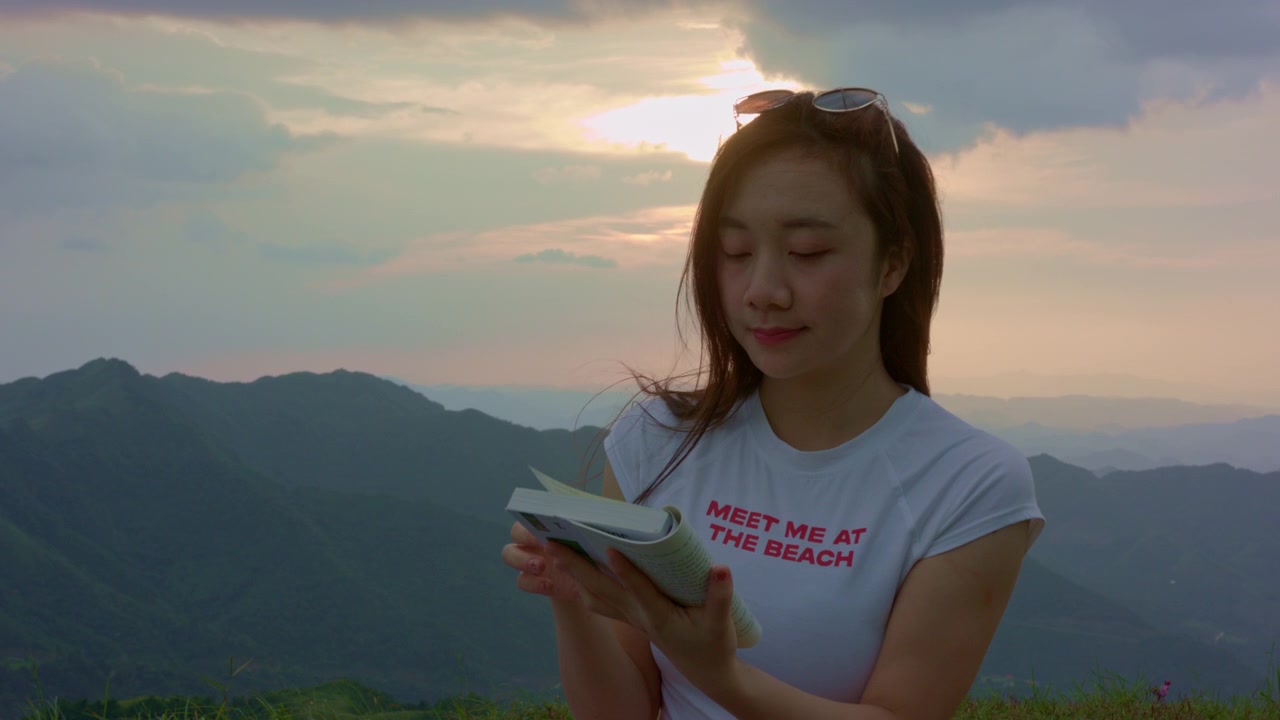 Wanita muda membaca buku di luar ruangan saat matahari terbenam, dikelilingi puncak gunung berlapis dan cahaya latar hangat, menciptakan suasana perjalanan dan gaya hidup yang damai dari sudut pandang yang indah