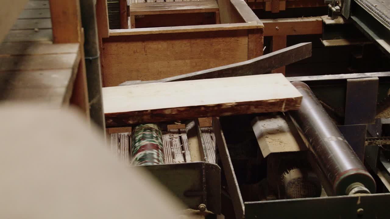 A close-up of industrial machinery in a sawmill. A powerful, serrated metal roller feeds a raw, live edge wood plank forward for processing, showcasing the timber industry