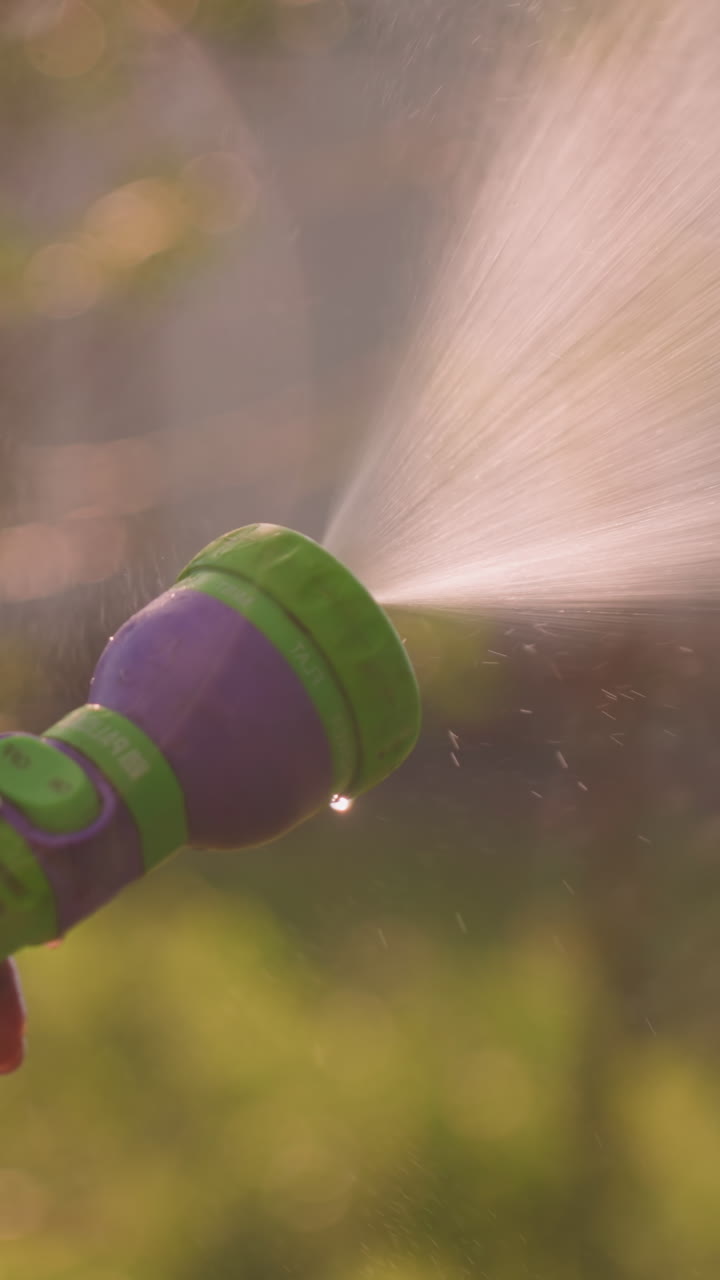 Close-up of someone holding water hose spraying water outdoors, creating refreshing mist effect, with blurred background of greenery and building in soft focus