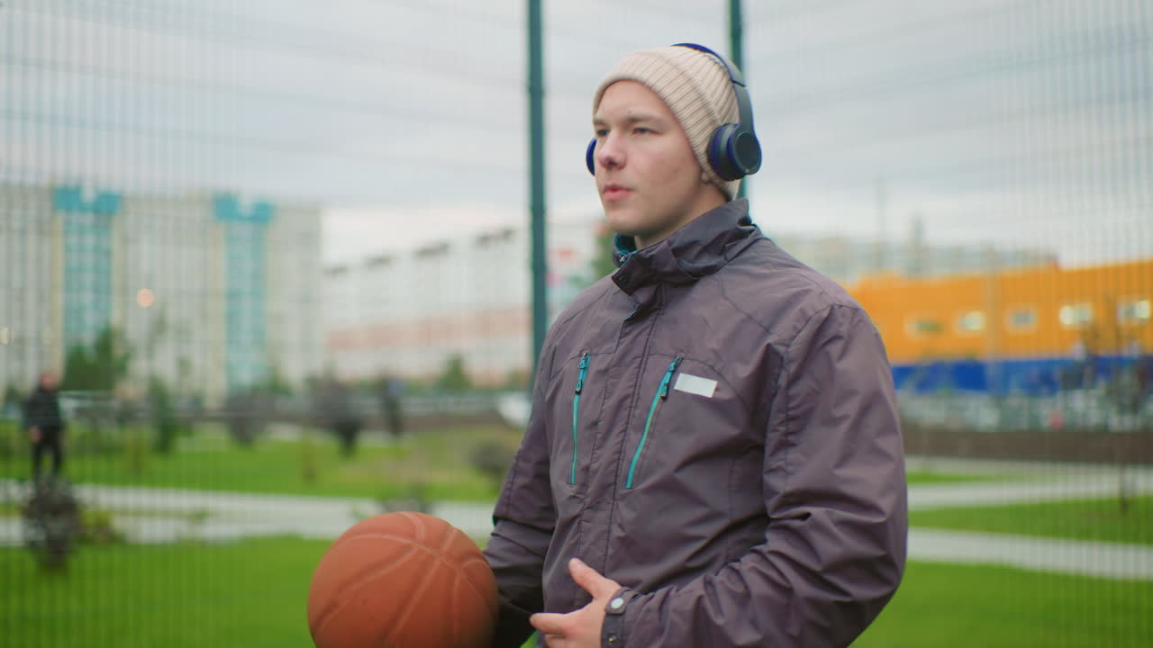 Ein Mann mit Basketball auf dem Feld, der Kopfhörer und eine Mütze trägt, steht an einem Metallzaun in einem bewölkten Stadtpark, hält den Ball, während er Musik hört und sich auf das Training vorbereitet – ein ruhiger, konzentrierter Gesichtsausdruck.
