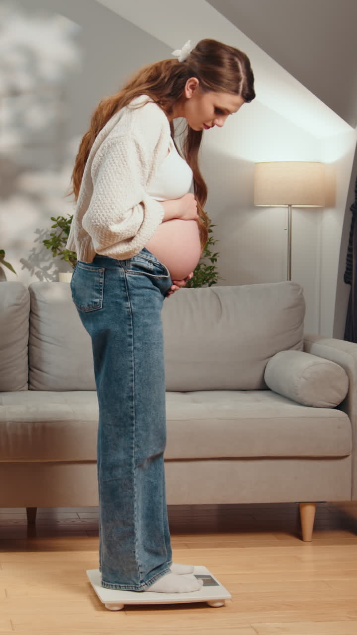 Pregnant woman stands on scale checking weight gain during pregnancy indoor home setting with sofa