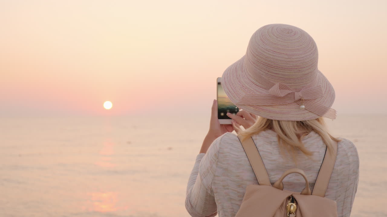 una chica rubia con sombrero y mochila se encuentra con el comienzo de un nuevo día junto al mar y toma ap
