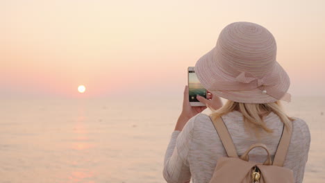 una chica rubia con sombrero y mochila se encuentra con el comienzo de un nuevo día junto al mar y toma ap