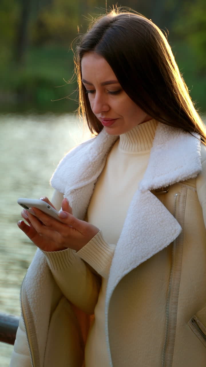 Relaxed serious lady standing on the bridge over the river. Attractive lady using the phone holding in her hand. Blurred nature backdrop.