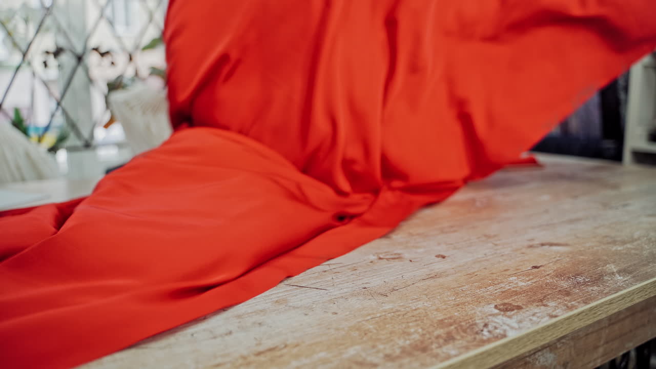 Seamstress laying out beautiful red cloth on a wooden table. Work place table of tailor in workshop and a big piece of red fabric indoors.
