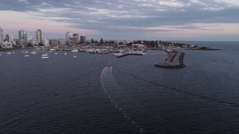 cinematic aerial circling shot of red fishing motorboat entering the city harbor of punta del este in uruguay with skyline and skyscrapers in background