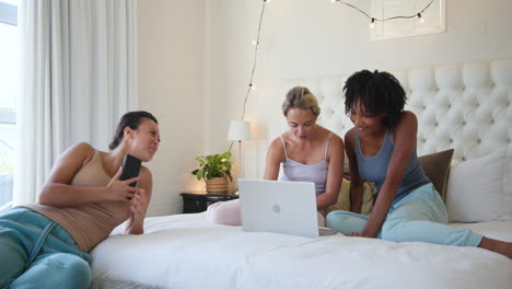 diverse female friends relaxing on bed, using laptop and chatting happily together