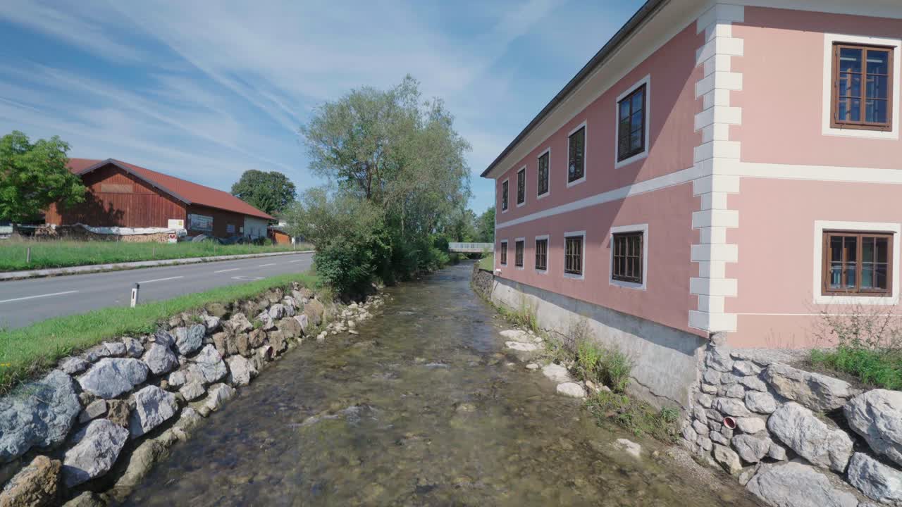 Halbnahe Einstellung eines kleinen Flusses, der zwischen einer Straße und einem rosa Haus in Österreich fließt.