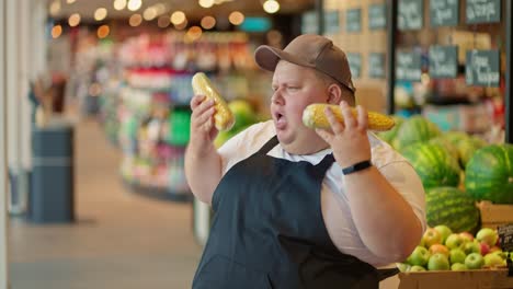 A-cheerful-overweight-male-supermarket-worker-in-a-white-T-shirt-and-a-black-apron-in-a-gray-cap-dances-along-with-heads-of-corn-and-uses-them-as-a-microphone-during-his-fun-at-work-against-the-background-of-counters-with-vegetables-and-fruits-in-a-supermarket