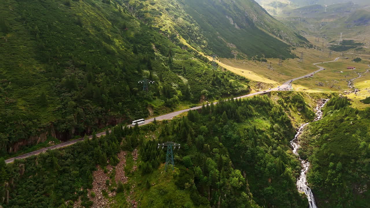 White bus rides by the highway in the beautiful green mountains. Drone footage over the narrow river flowing in the gulch. Romania nature